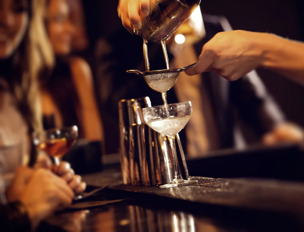 Bartender pouring a cocktail from a shaker into a coupe glass.