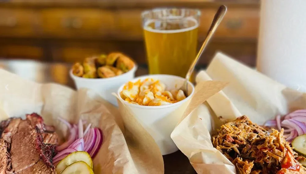 Barbecue spread with sliced brisket, pulled pork, macaroni, pickles, and beer.