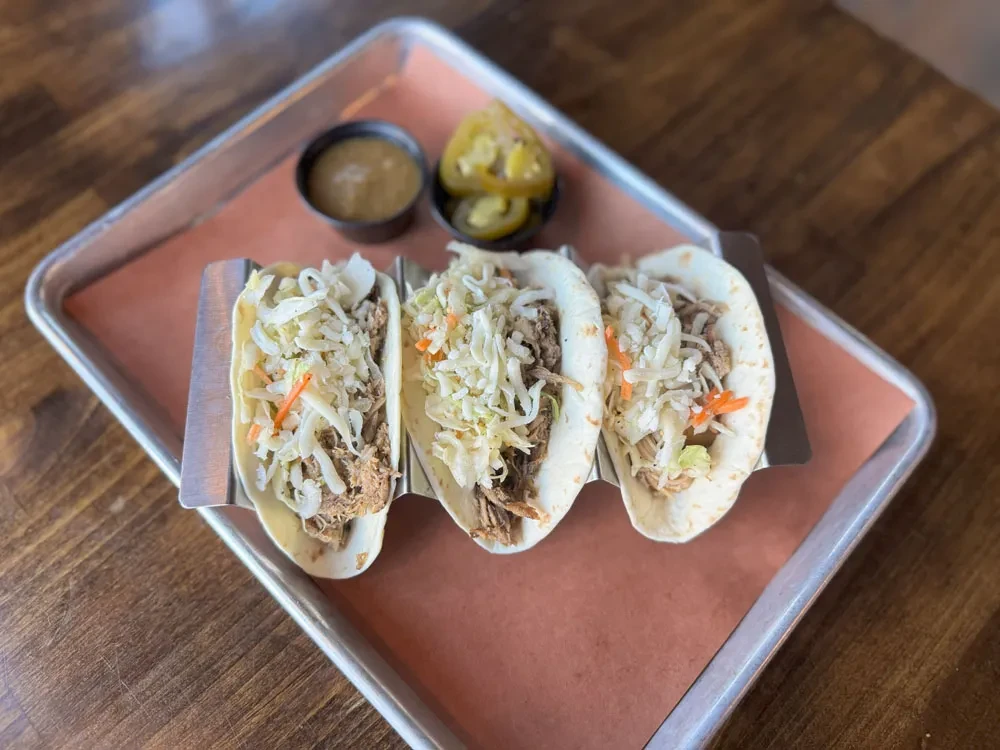 Three meat tacos with coleslaw, sauce, and pickled peppers on a wooden table.
