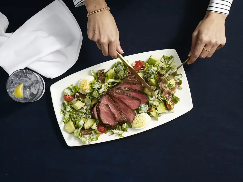 Hands with gold cutlery cutting medium-rare steak salad.