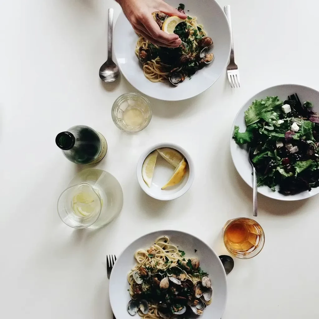 Overhead flat-lay of spaghetti and clams, salad; hand squeezes lemon.