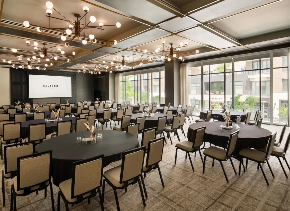 Modern banquet hall with round tables, chandeliers, city view, and 'HALCYON' screen.