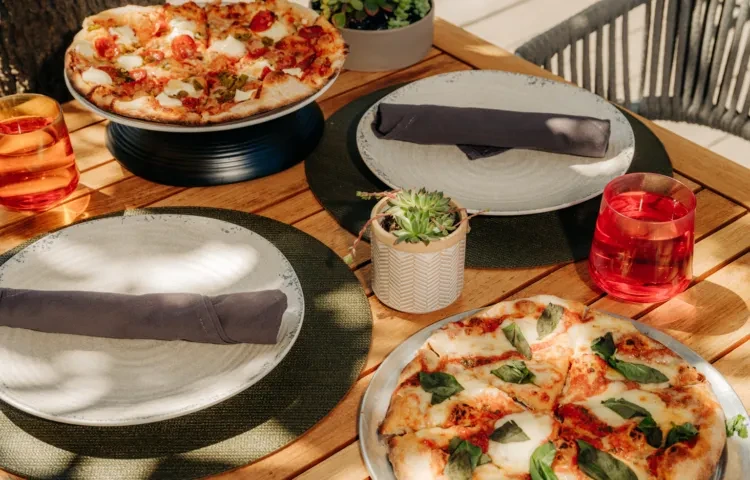 Overhead of two pizzas and drinks on a sunlit outdoor wooden table.
