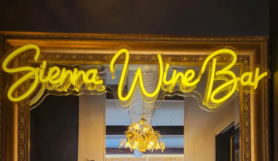 Yellow neon 'Sienna Wine Bar' sign reflected in an ornate gold mirror.
