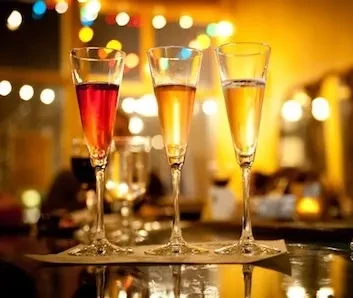Three glasses with red, amber, yellow drinks, against blurred lights.