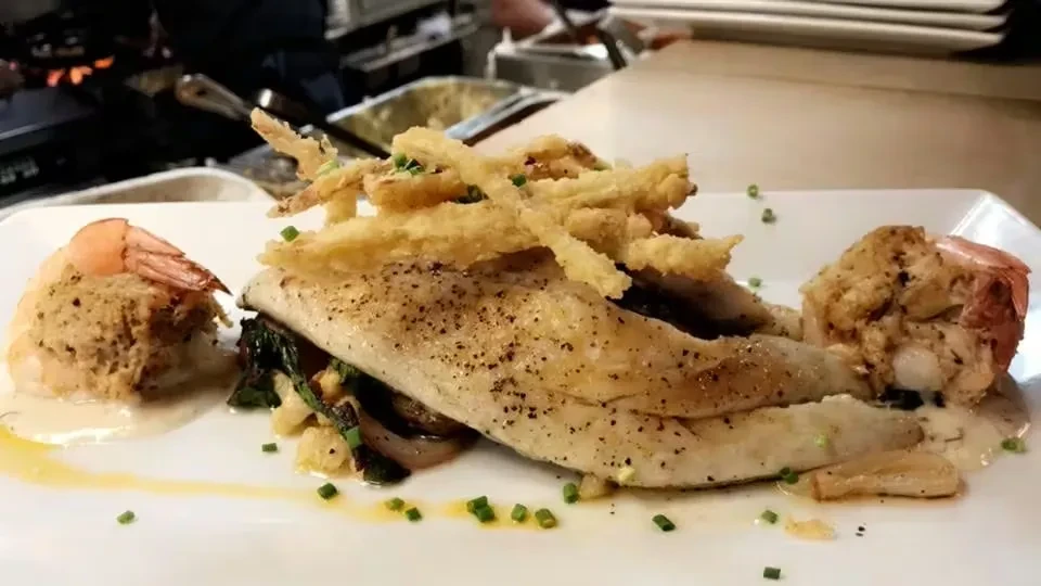 Plated fish fillet, shrimp, crispy fried strips, greens, and cream sauce.