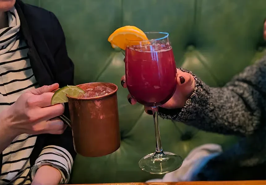 Two hands toast cocktails: a copper mug and a wine glass.