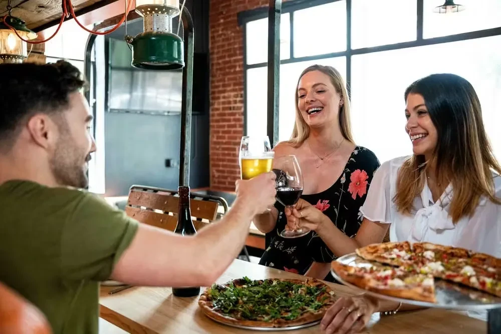 Three people joyfully toast with drinks and pizzas in a restaurant.