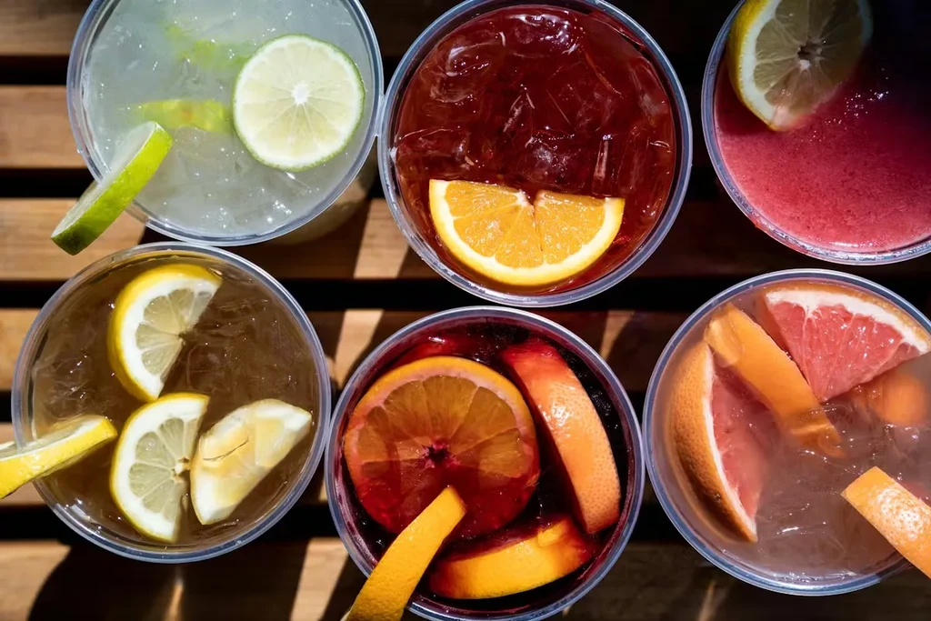 Six colorful fruit-garnished drinks on a wooden slatted surface.