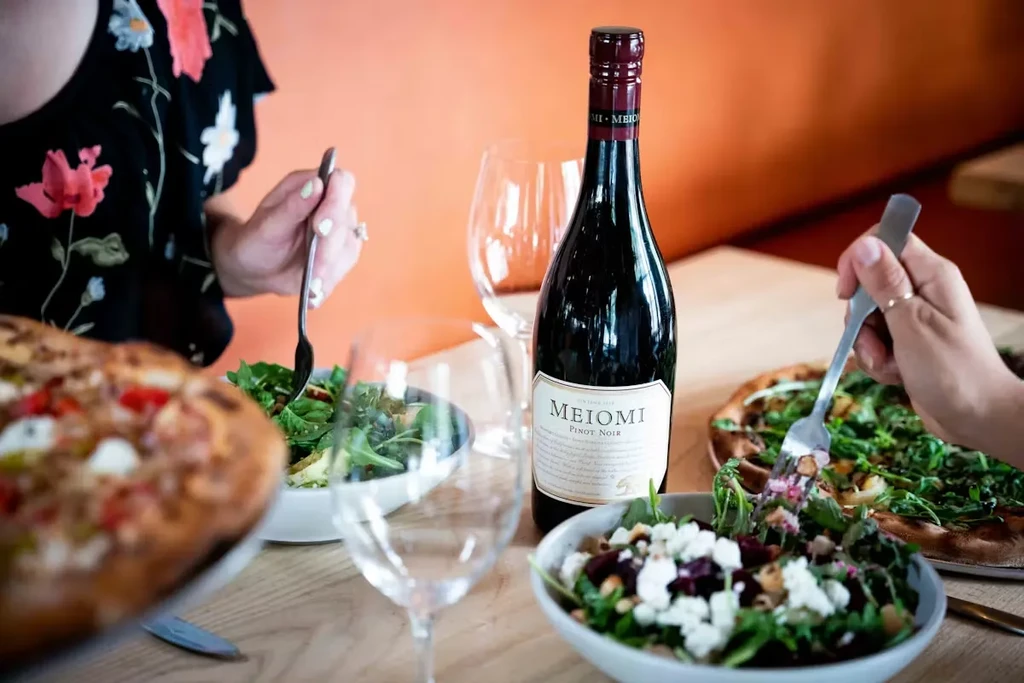 Dining table with salads, pizzas, Meiomi Pinot Noir, and people's hands.
