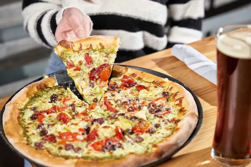 Hand lifts pizza slice with pesto, tomatoes, olives; beer nearby.