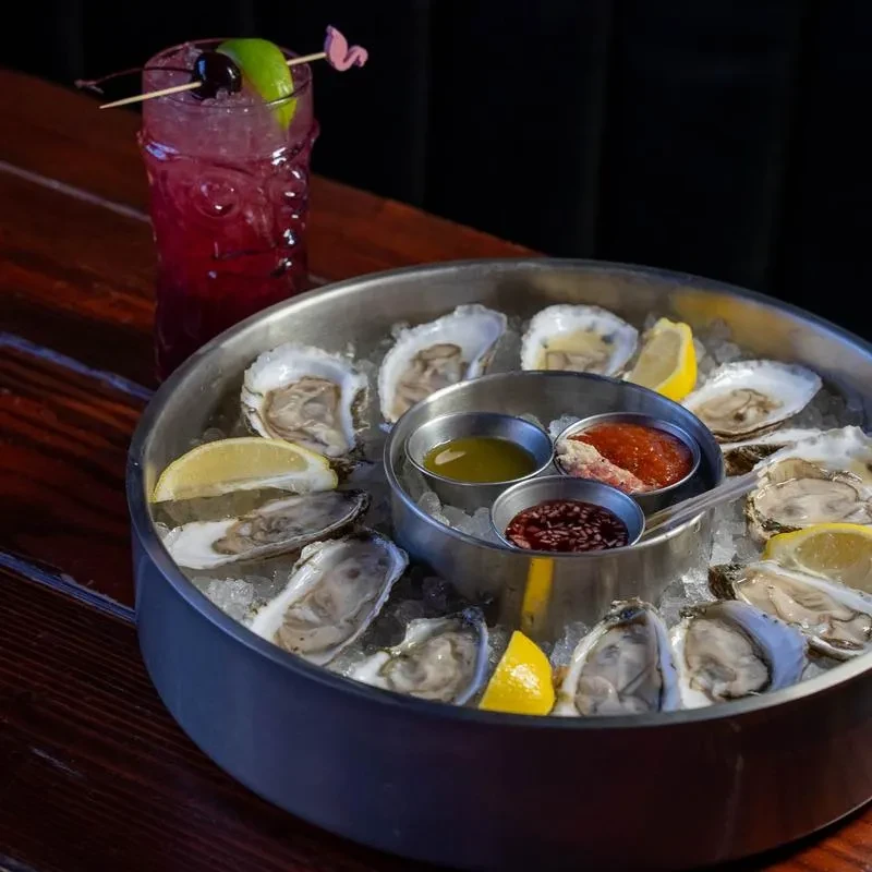 Raw oyster platter with lemon, sauces, and a purple cocktail.
