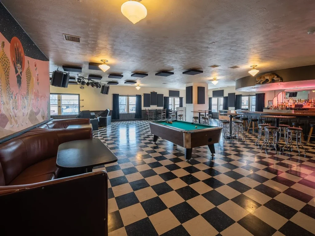 Empty bar interior, checkered floor, stage, pool table, full bar.