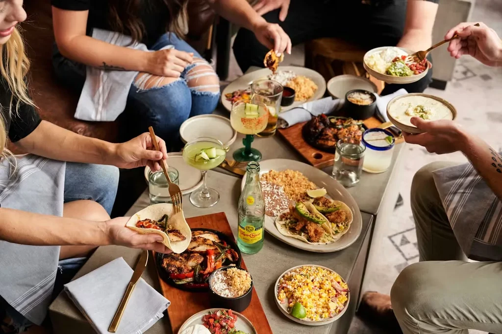 Communal table with tacos, guacamole, drinks, and interacting hands.