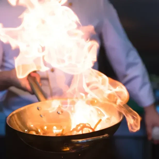 Chef's hands flambéing food in a pan, bright flames erupting.