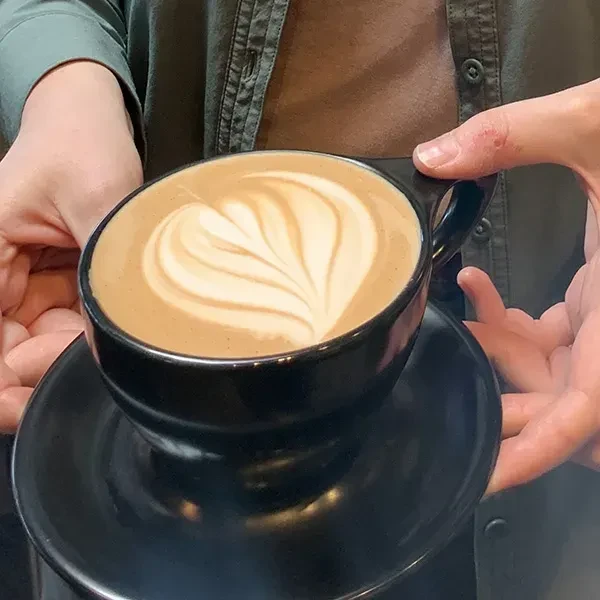 Hands hold a black ceramic latte with heart-shaped foam art.