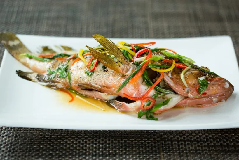 Whole reddish-pink fish with colorful vegetables on a white plate.