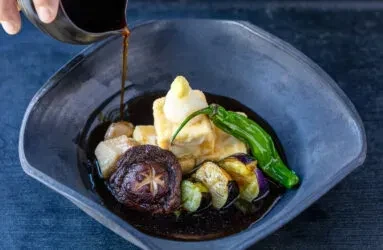 Japanese tofu dish with shiitake, eggplant, pepper, sauce pouring in dark bowl.
