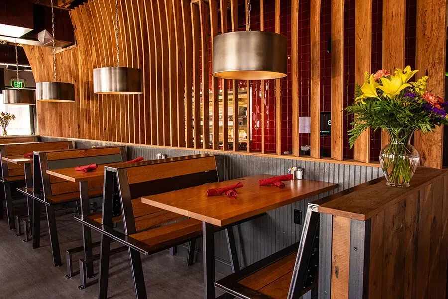 Empty restaurant with dark wood booths, slatted walls, pendant lights, and flowers.