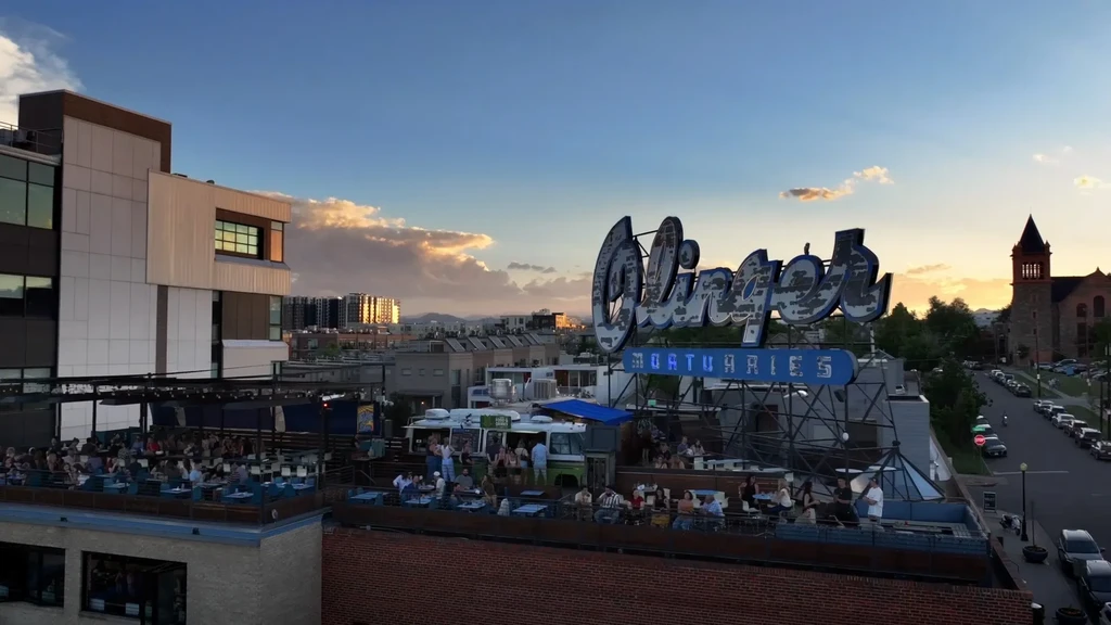 Rooftop bar at sunset with patrons and "OLIVE MORTUARIES" neon sign.