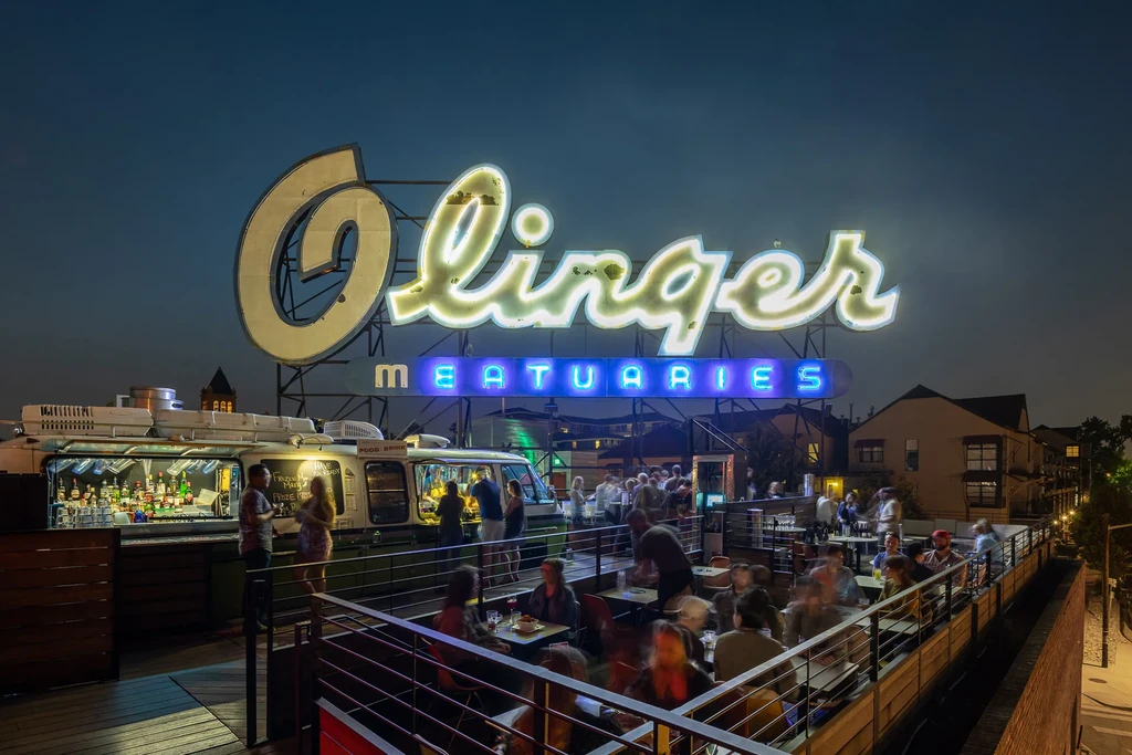 Rooftop bar with "Olinger MEATUARIES" neon sign and vehicle-themed bar.