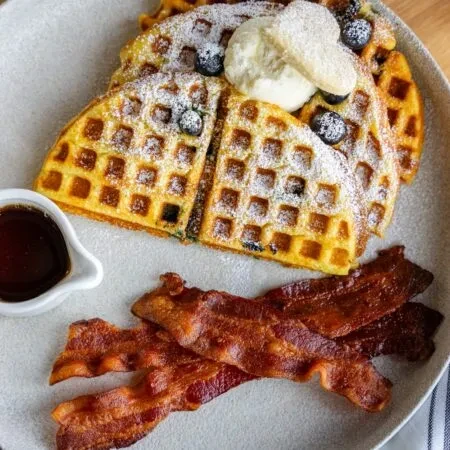 Plate of waffles with blueberries, cream, bacon, and syrup.