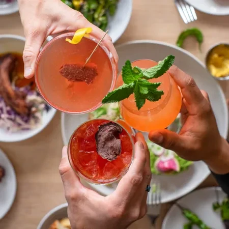 Three hands holding colorful cocktails over a restaurant table with food.