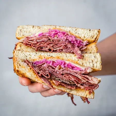Hand holding a large pastrami, cheese, and sauerkraut sandwich.
