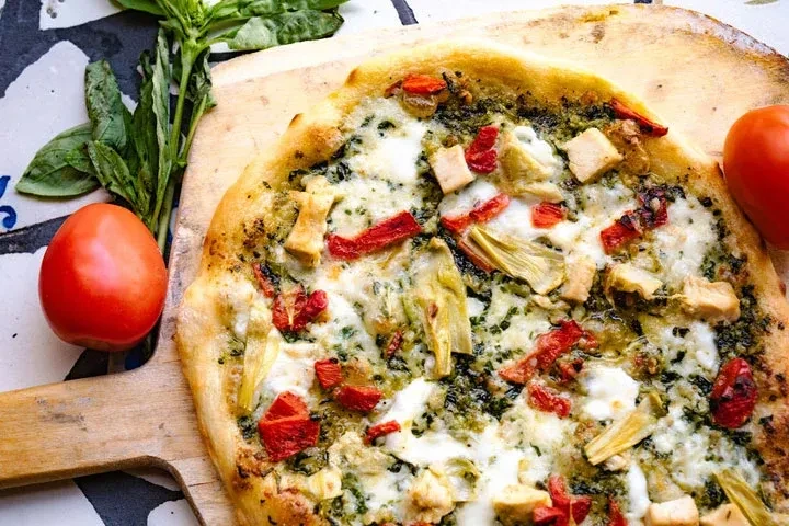 Overhead: Pesto chicken pizza with artichokes, red ingredients, basil, tomatoes.