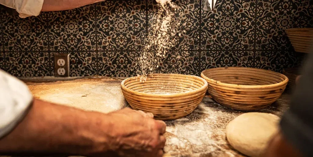 Baker sifts flour into a round proofing basket on a floured surface.