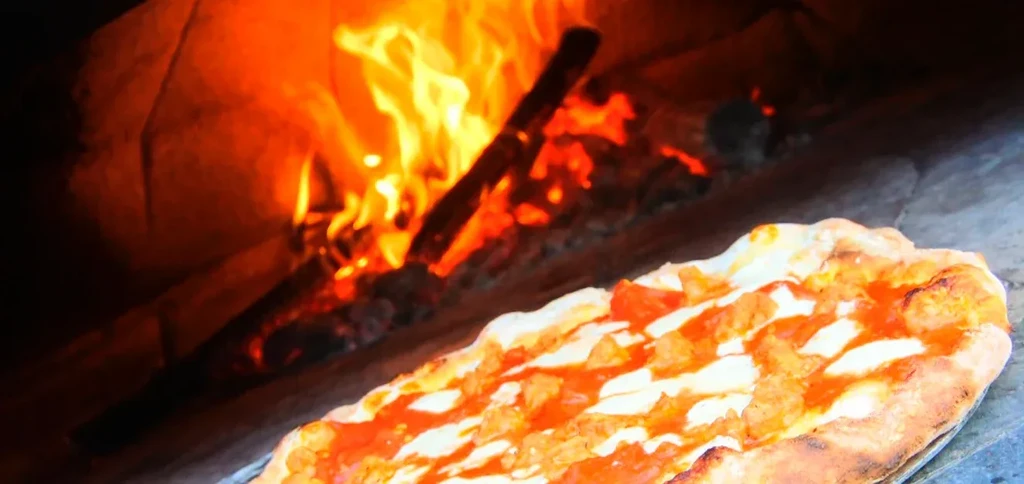 Pizza on peel in front of flaming wood-fired oven.