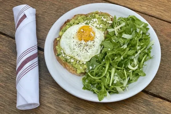 Avocado toast with fried egg, crumbled cheese, and green salad on wood.