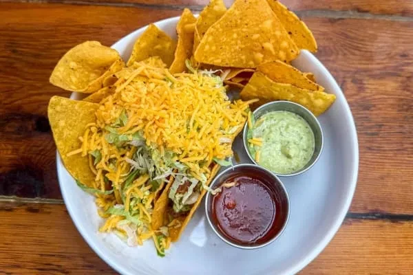 Top-down view of cheesy tacos, chips, guacamole, salsa on wooden table.