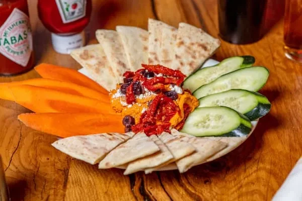 Platter with reddish-orange dip, sun-dried tomatoes, pita, carrots, and cucumbers.