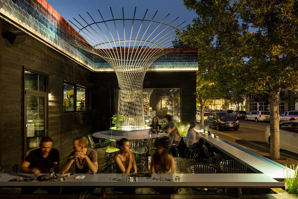 Modern restaurant exterior at night with colorful frieze and lit metal sculpture.