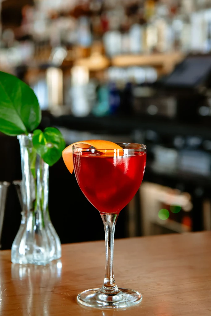 Deep red cocktail with orange zest on a wooden bar counter.