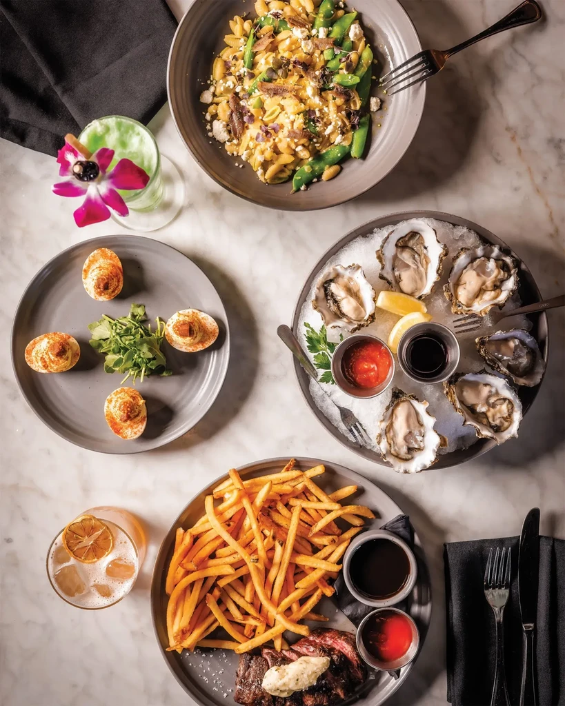 Marble table with pasta, steak, oysters, deviled eggs, and two cocktails.