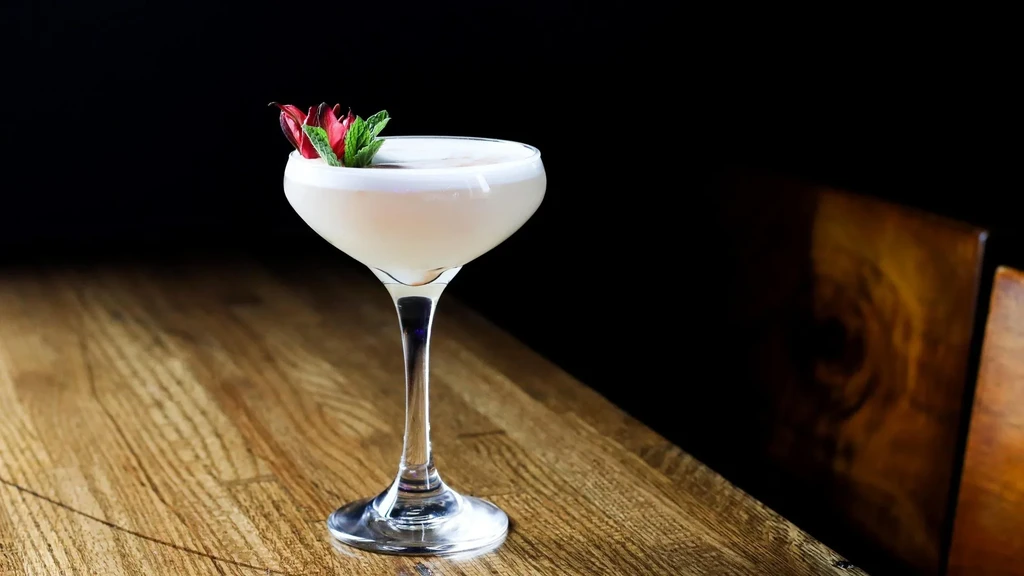Creamy white cocktail in coupe glass, garnished with mint and red petals.