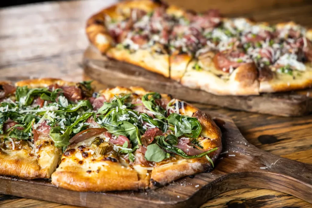 Sliced oblong pizza with meat, arugula, cheese; another in background.