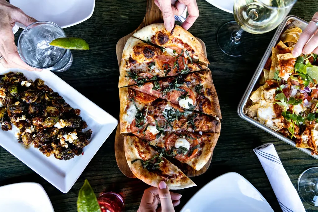 Overhead view: Hands sharing pizza, loaded nachos, and roasted Brussels sprouts.