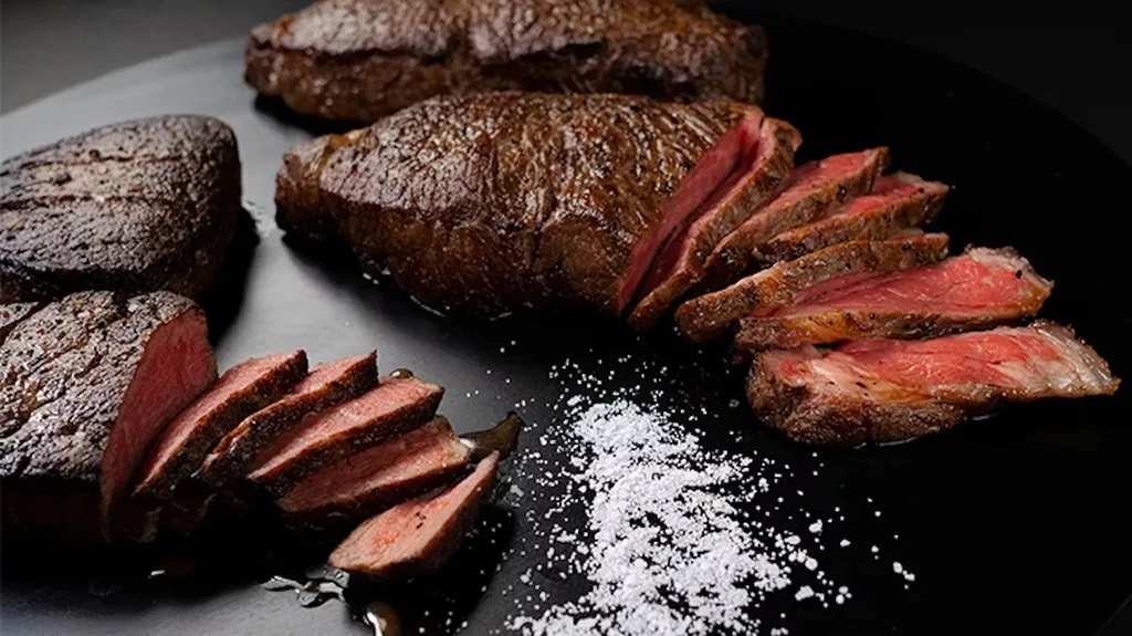 Three medium-rare seared steaks on a dark surface with salt.