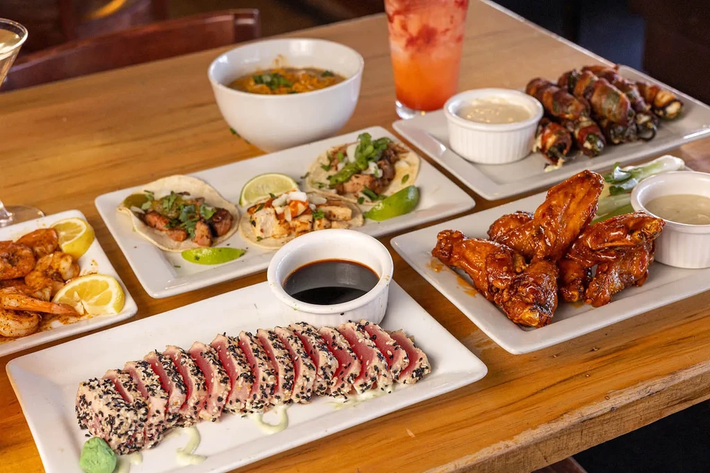 Table with seared tuna, shrimp, tacos, chicken wings, and beverages.