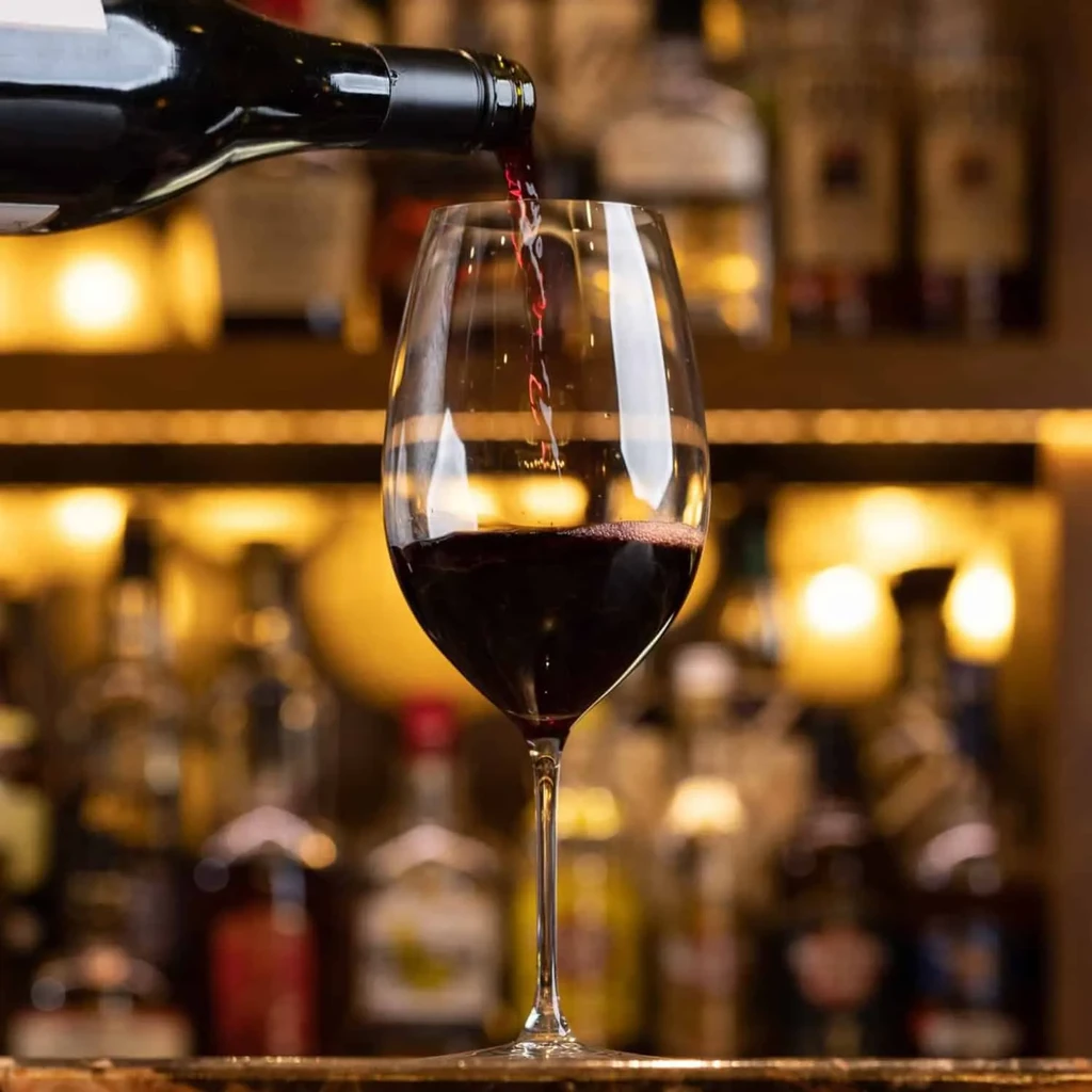 Red wine pours from bottle into a glass, blurred bar background.