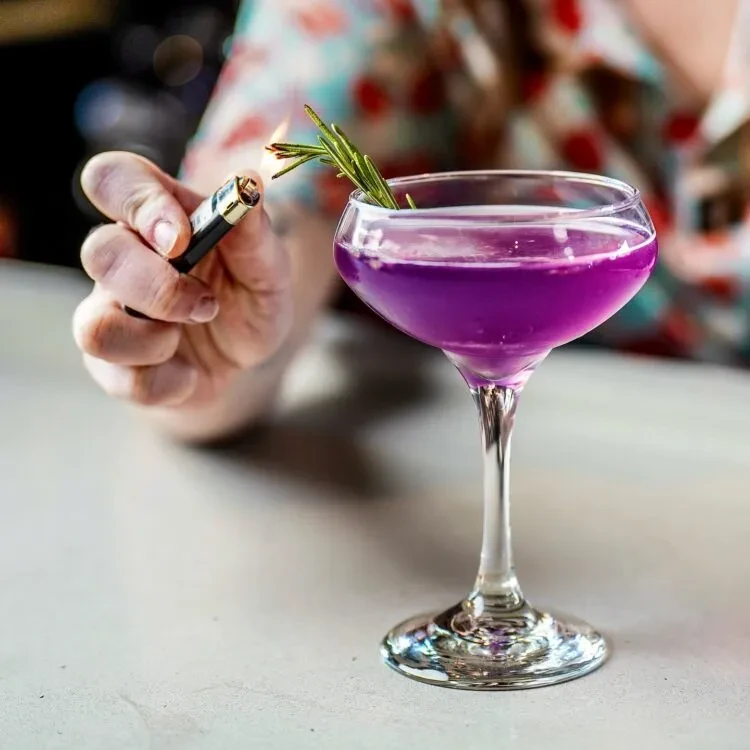 Purple cocktail in coupe glass with flaming rosemary garnish.