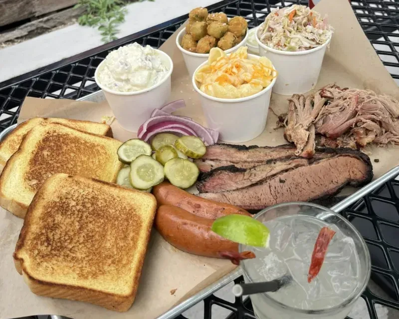 Barbecue platter with brisket, pulled pork, sausages, sides, and a cocktail.