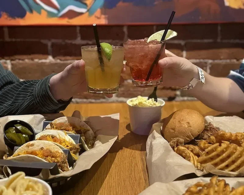 Two hands toast cocktails over tacos, BBQ sandwich, and fries.