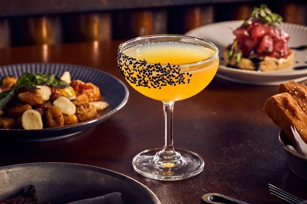 Yellow cocktail in coupe, black sesame rim, surrounded by blurred dishes.