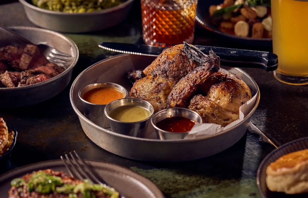 Rustic table with roasted chicken, grilled steak, sauces, and drinks.