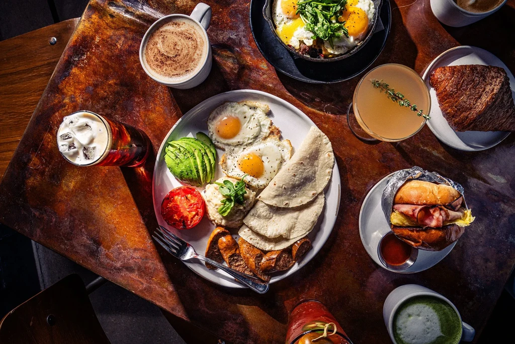 Diverse breakfast/brunch spread with eggs, pastries, and drinks on a dark table.