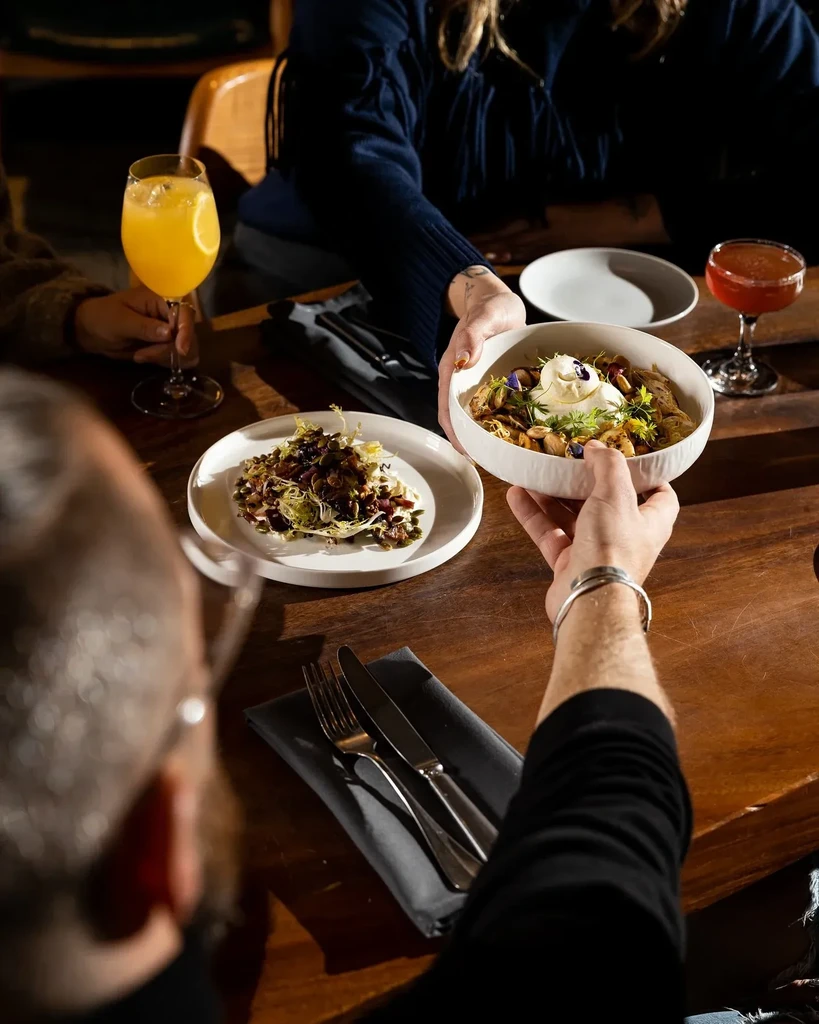 Hands pass a garnished pasta bowl, with a salad and cocktails.